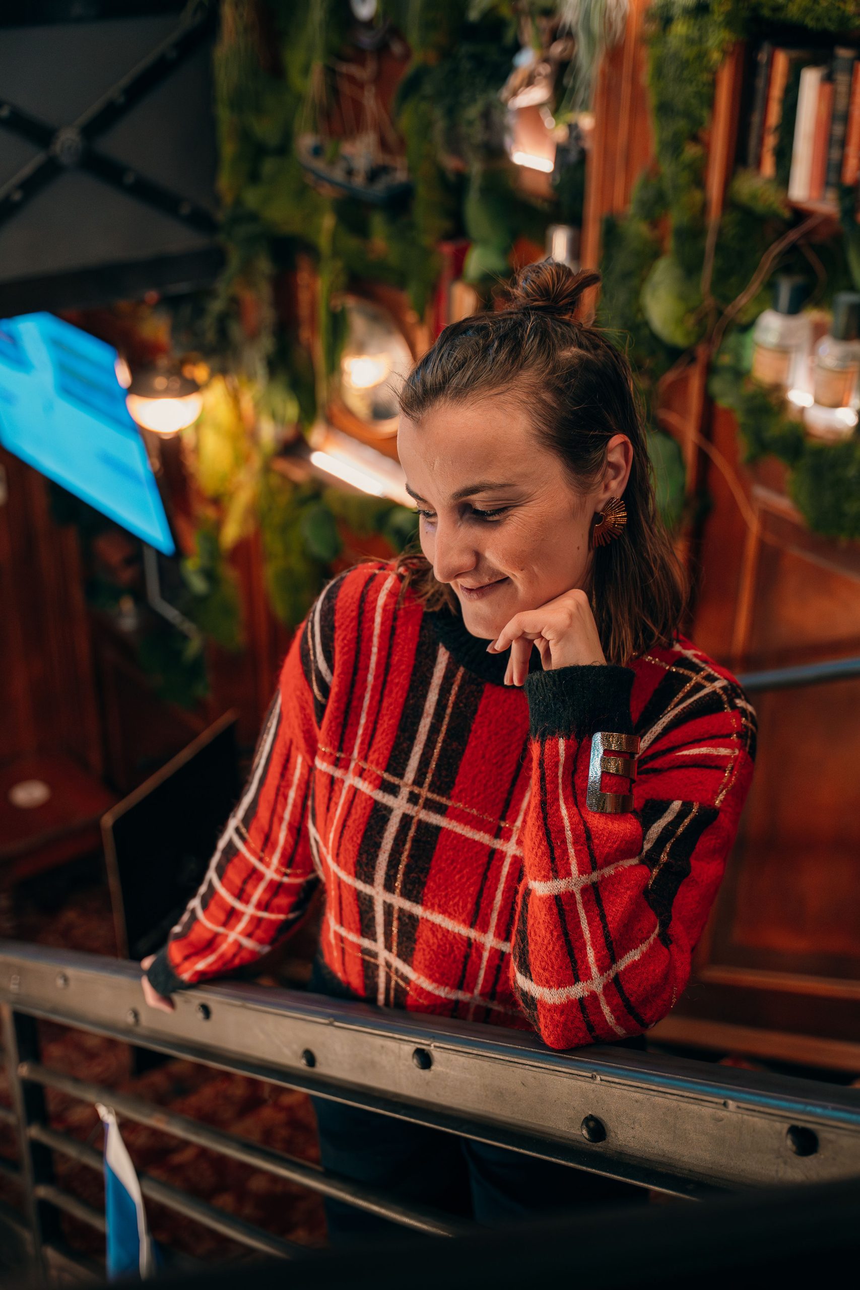 Portrait en pull à carreaux rouge, dans un décor chaleureux de livres et de végétation.