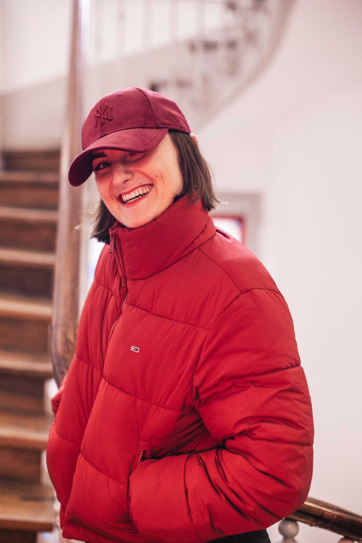 Un grand sourire, une veste rouge et une casquette bordeaux, près d'un escalier.
